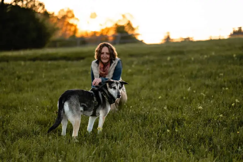 Almut kniet im Abendlicht auf einer Wiese neben ihrem Hund