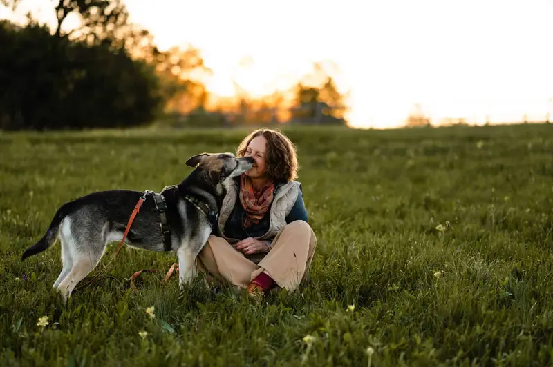 Almut sitzt lachend auf einer Wiese, ihr Hund schleckt ihr übers Gesicht