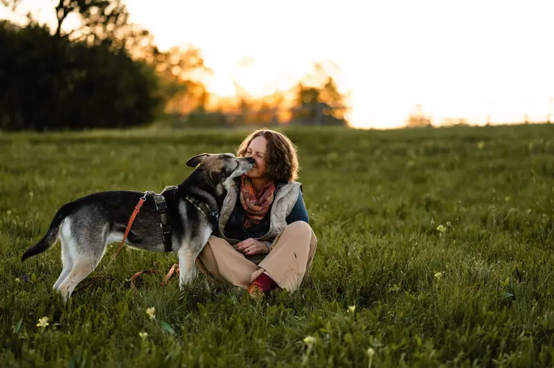 Almut sitzt im Abendlicht auf einer Wiese, ihr Hund gibt ihr einen Kuss
